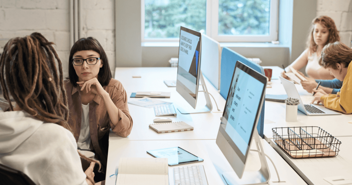 Students collaborate at a bright shared desk with desktop computers and notebooks in a modern classroom setting.