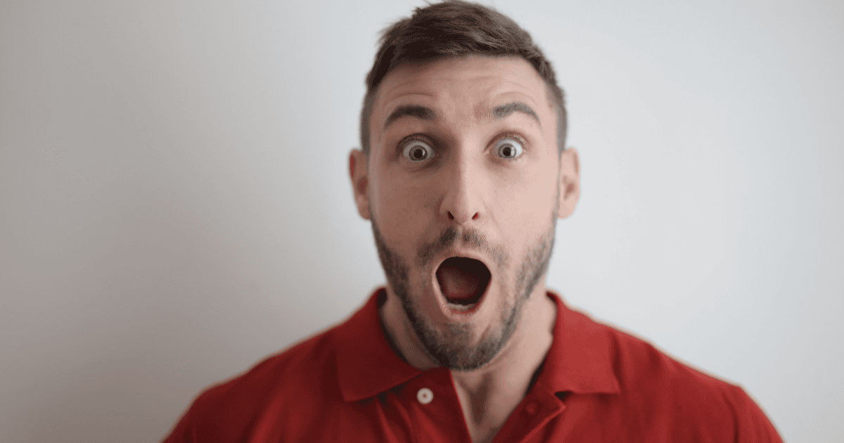 Man with short dark hair and a beard, wearing a red polo, eyes wide and mouth open in surprise.
